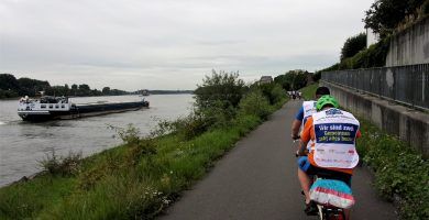 Auf dem Rhein ein Schiff, auf dem Fahrradweg daneben Teilnehmer des Tandem-Camps